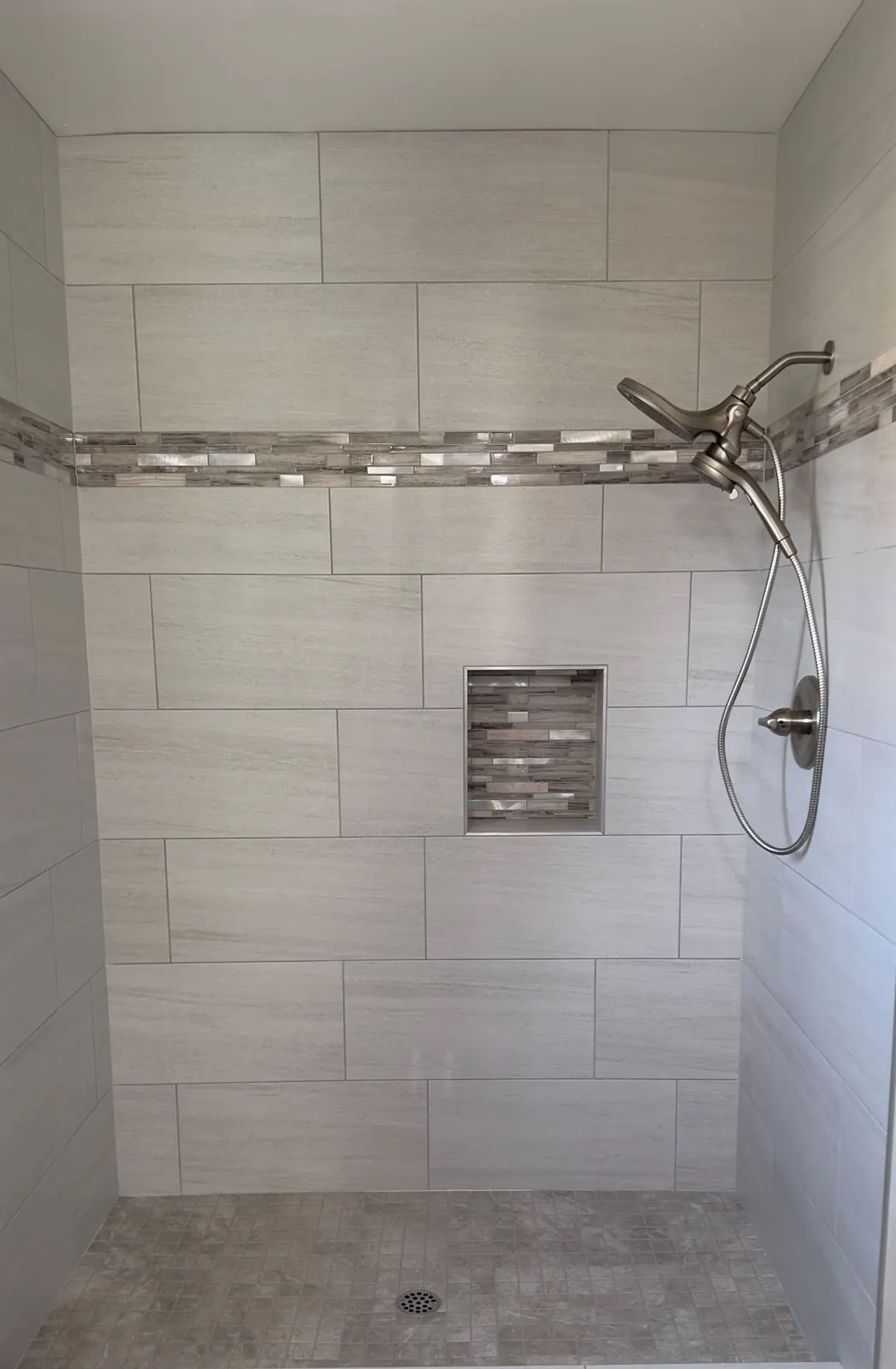 Shower with large light grey wall tiles, a metal and glass mosaic band and matching niche, and a small square mosaic floor